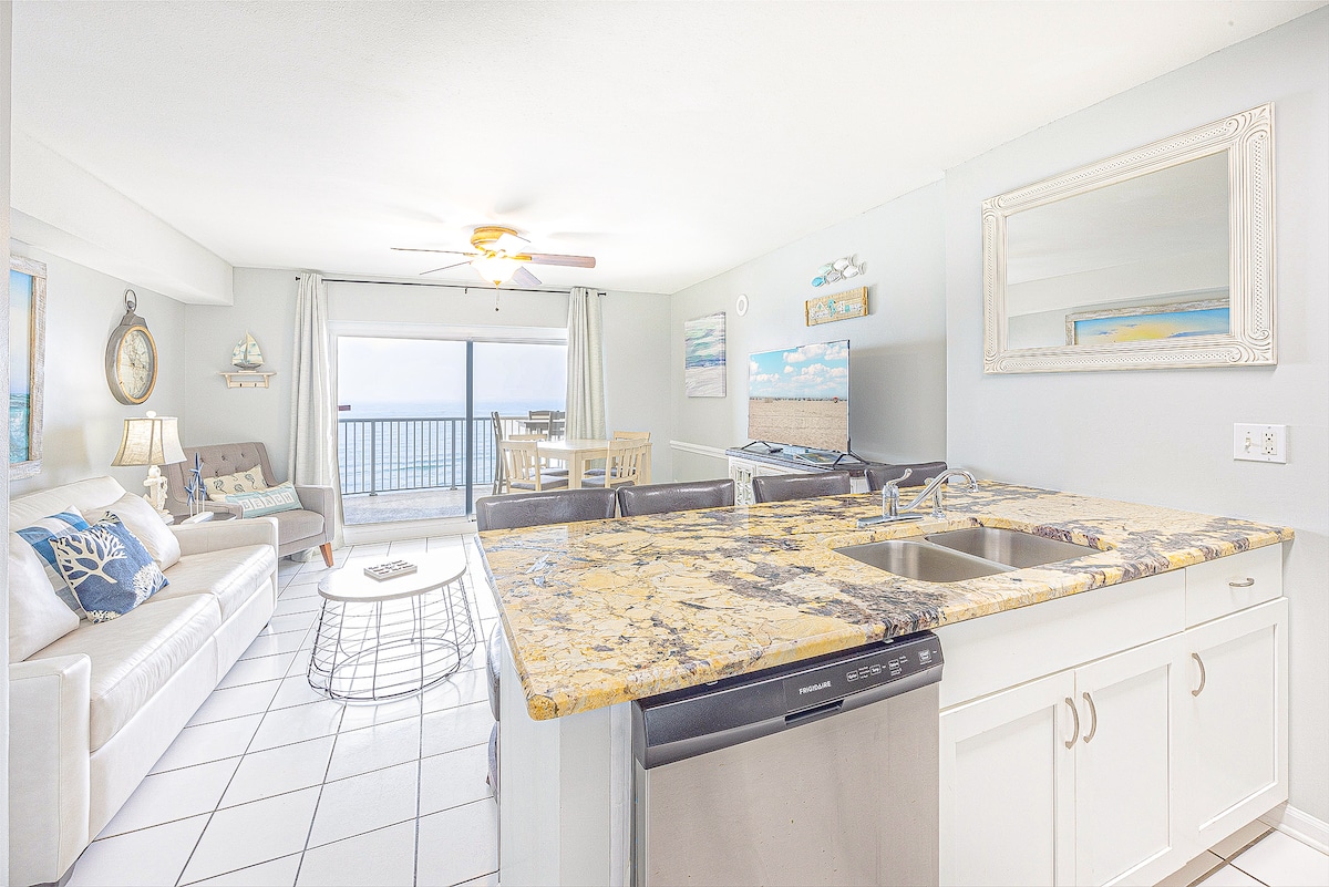 An open concept living area features a granite-topped kitchen island with double sinks, overlooking a cozy seating area. A ceiling fan circulates air while a large window showcases a view of the Gulf. Light-colored furnishings and neutral tones invite relaxation in this space.