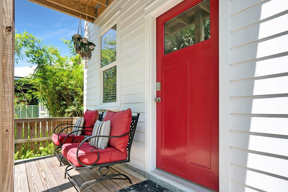 A welcoming entryway is presented with a bright red door and two comfortable chairs adorned with striped cushions. A textured welcome mat lies beneath, and vibrant greenery is visible, adding a refreshing touch to the porch space.