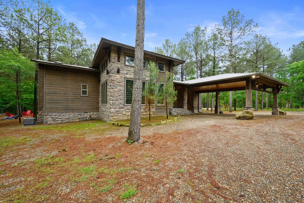 A contemporary cabin is showcased amidst a natural setting, featuring a blend of wooden and stone elements. The structure is complemented by tall pine trees and a gravel driveway. A spacious covered carport is also visible, offering protection from the elements.