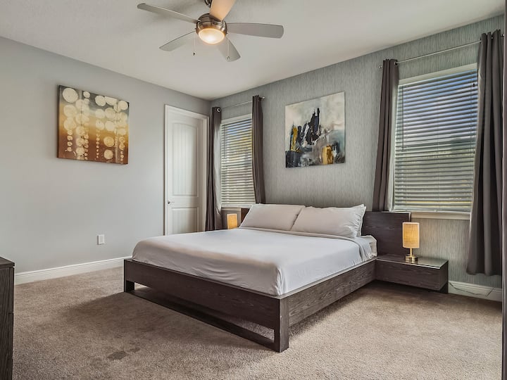 - Брачна спална соба со едноставен модерен декор и телевизор  
- Природна светлина низ големи прозорци со завеси  
- Удобно место за одмор со мирен семеен амбиент