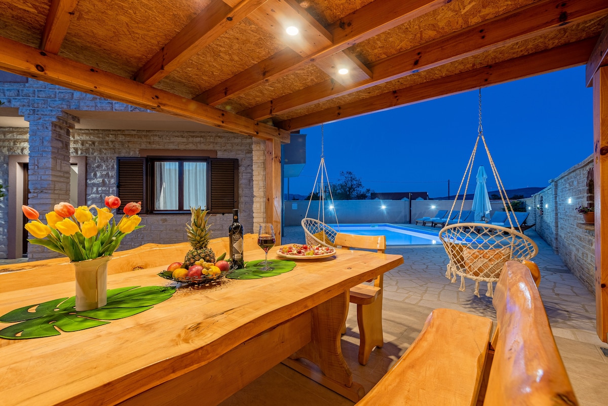An outdoor dining area features a large wooden table set with a platter of fruit. Bright tulips in a vase add a touch of color. Swing chairs hang from the ceiling, and a swimming pool is visible in the background under a twilight sky.