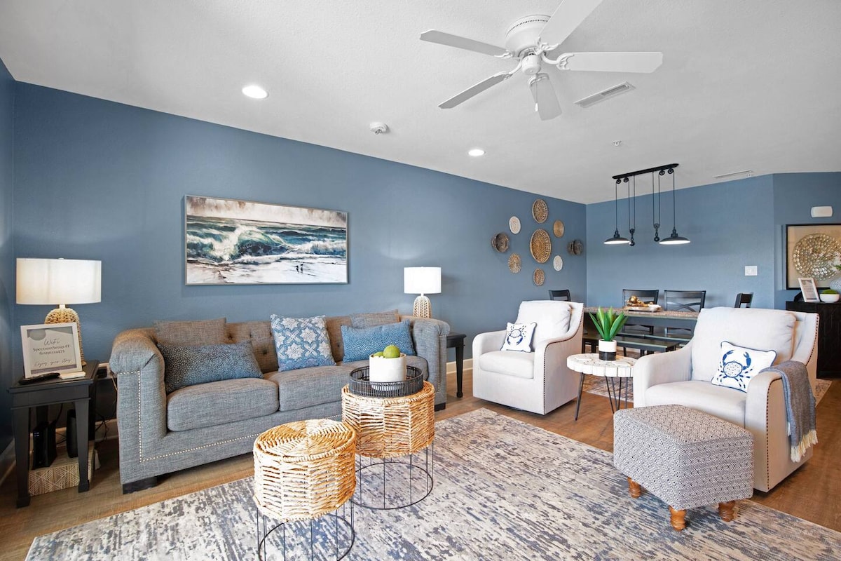 A modern living area is presented with a gray sofa and two white armchairs. Distinctive round coffee tables sit on a light-colored rug, and decorative wall clocks adorn the blue walls. Natural light is illuminated from overhead fixtures, creating a welcoming space.