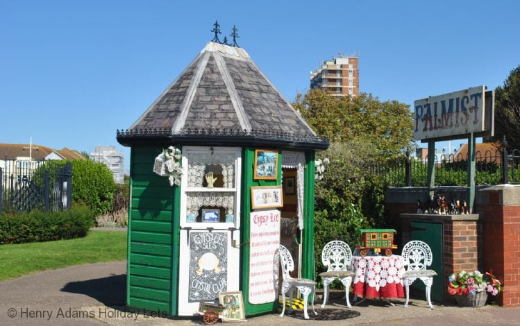 A small, round building with a thatched roof features a welcoming façade painted in green. Displayed are signs for palm reading. Two white chairs with intricate designs sit outside, accompanied by a table adorned with lace. Lush greenery and urban buildings are visible in the background.