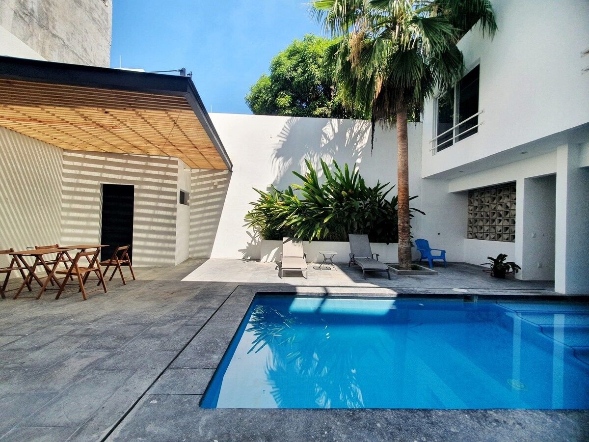A shared pool area features a clear blue pool surrounded by lounge chairs and tropical plants. Shade is provided by an overhead structure, with a small table and chairs positioned nearby, creating a relaxing outdoor space.