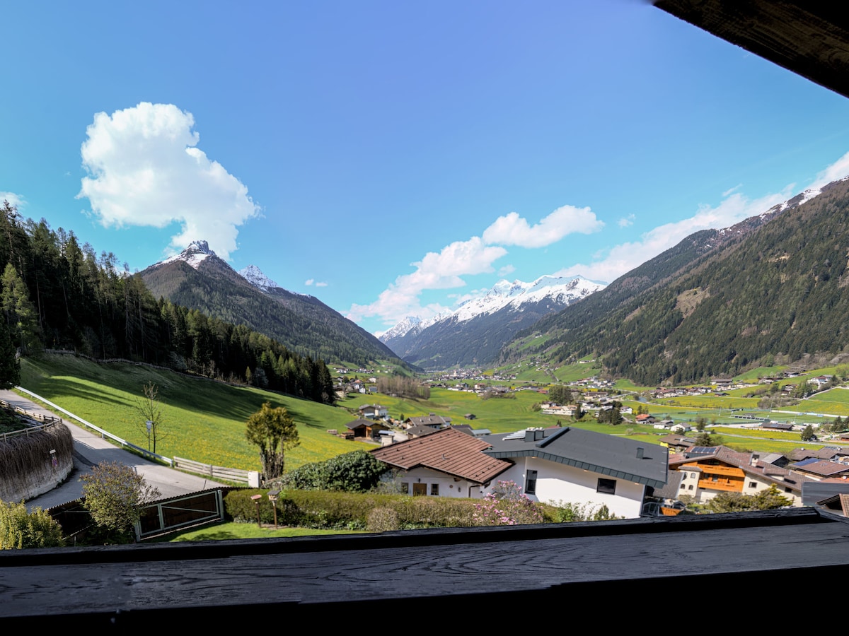 A panoramic view of the lush green valley is captured, bordered by distant mountains and dotted with quaint village homes. The sky is bright with fluffy white clouds, creating a serene backdrop to the natural landscape.