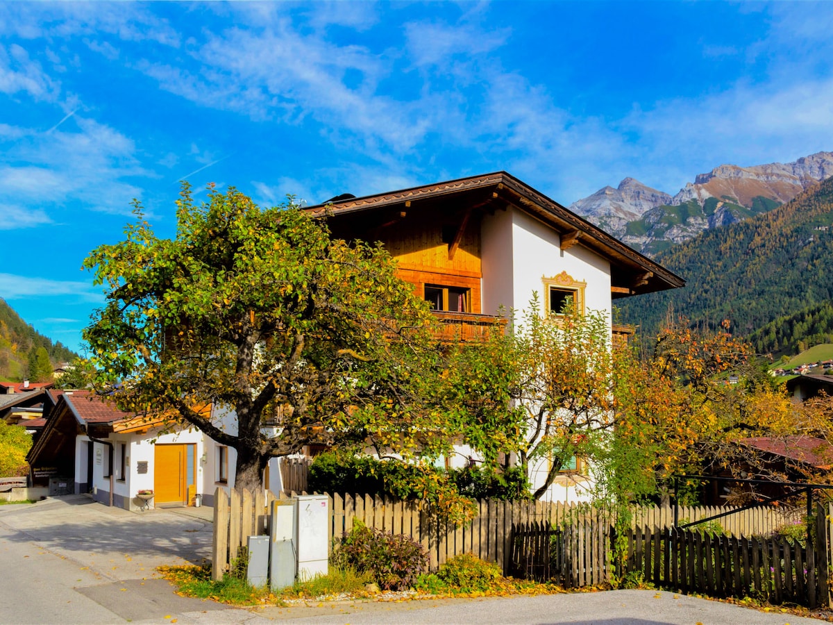 A charming two-story building is surrounded by trees and greenery, showcasing a wooden balcony on the upper floor. The structure features a blend of natural wood and white plaster, set against a picturesque mountain backdrop under a clear blue sky.