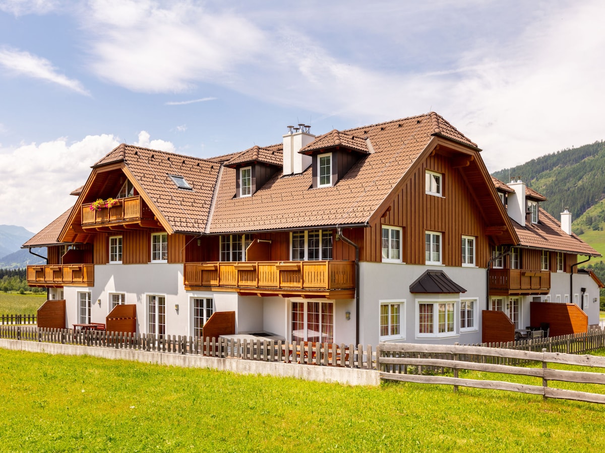 The exterior of a modern chalet is presented, featuring a combination of wood and stucco finishes. Balconies are visible on the upper levels, and greenery surrounds the property. Large windows are framed by a scenic backdrop of rolling hills and a clear blue sky.