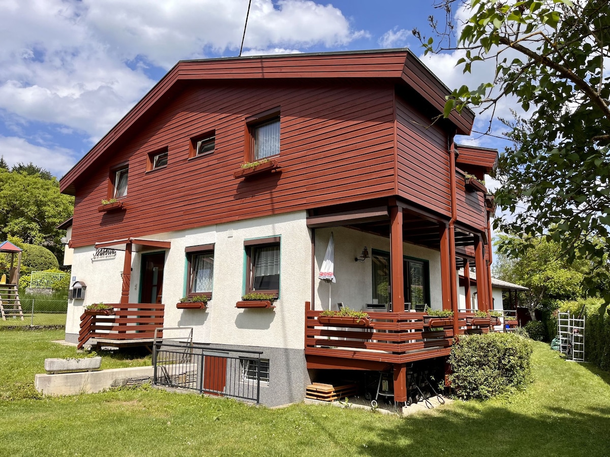 A two-story apartment house features a warm red exterior with wooden accents. The building is partially surrounded by greenery and has outdoor seating on the balconies. Large windows are adorned with flower boxes, providing a welcoming appearance.