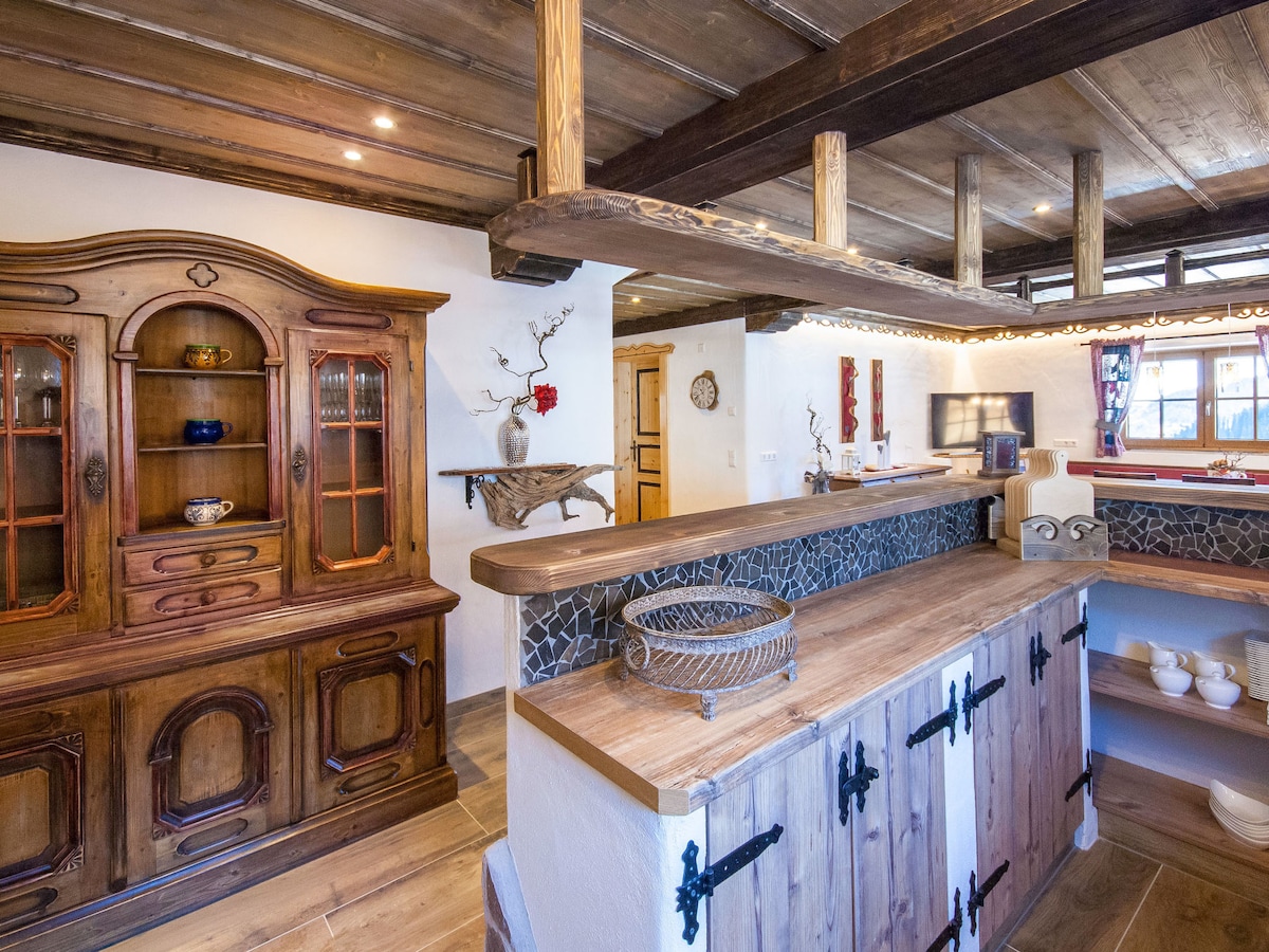 A rustic wooden cabinet with glass doors displays various dishware, complemented by a wooden countertop with a subtle texture. Natural light filters in through windows, illuminating the warm tones of the wooden beams and furniture throughout the inviting kitchen space.