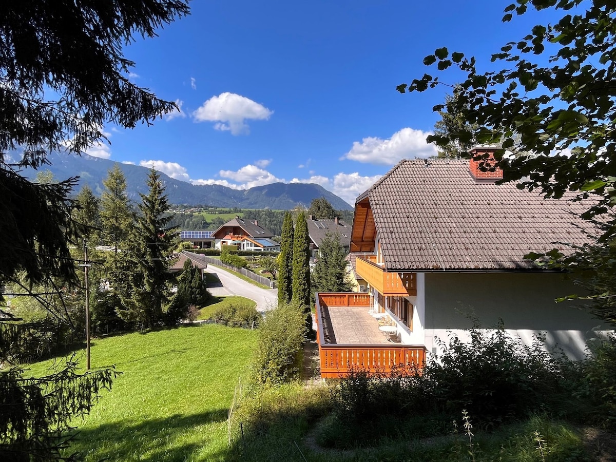 A scenic view captures the exterior of the two-family house surrounded by lush greenery and mountains. A spacious balcony is visible, offering a platform to enjoy the fresh air, while contrasting wooden accents highlight the home's architecture against the bright blue sky.