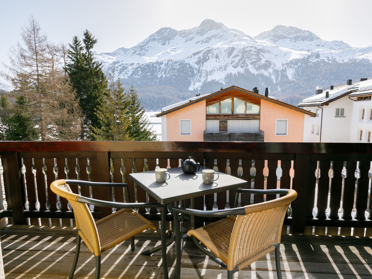 A balcony view is presented with two wicker chairs and a small table. The surrounding landscape features snow-capped mountains as a backdrop, with nearby chalet-style buildings visible. The area is enhanced by pine trees, providing a serene outdoor setting.