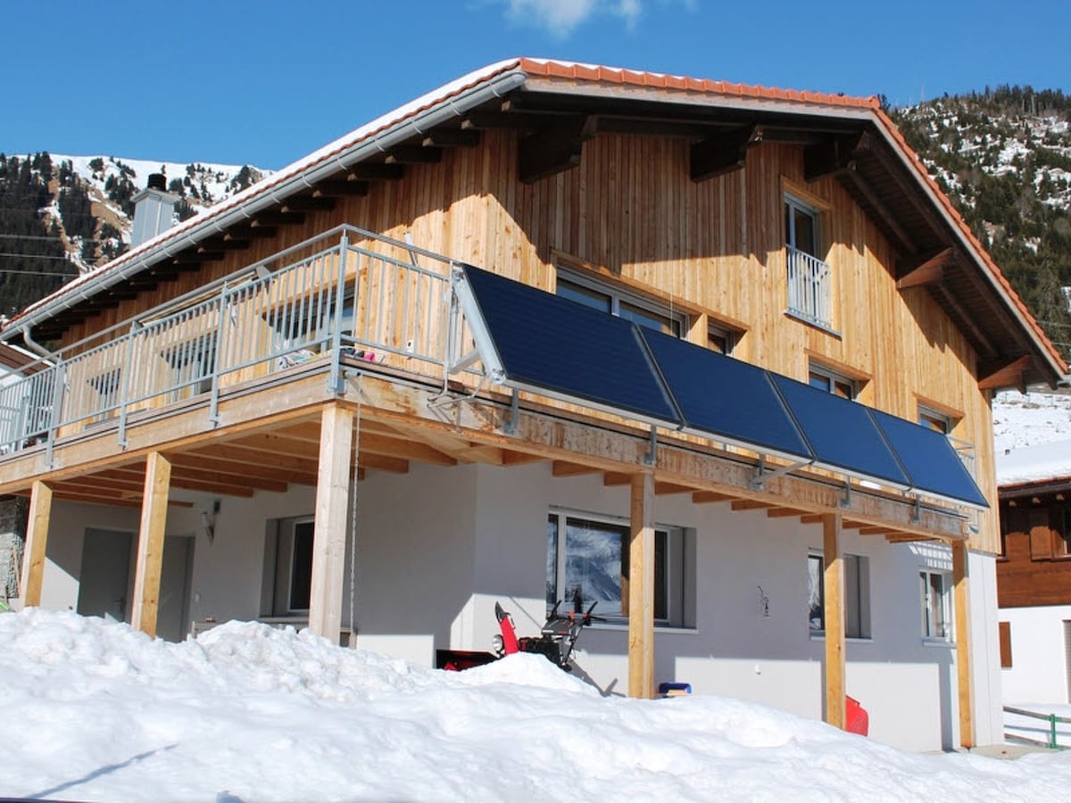 The exterior of a multi-family house features a wooden upper structure with a balcony, supported by wooden beams at the lower level. Solar panels are installed on the roof. Snow covers the ground, and the surrounding mountains can be seen in the background.