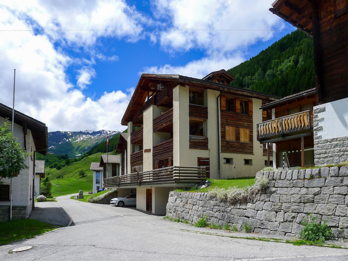 A three-story rustic apartment building is surrounded by lush green hills and mountains. The structure features multiple balconies with wooden railings and a stone wall lining the road. The pathway leads towards the building, under a partially cloudy blue sky.