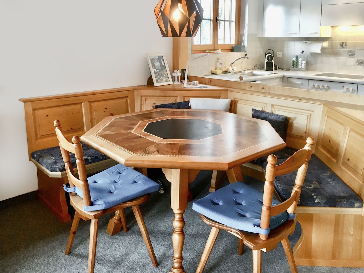 A wooden octagonal dining table is surrounded by four upholstered chairs, featuring a central black inlay. The seating area includes built-in benches adorned with blue cushions. Warm wood tones and a pendant light above enhance the welcoming space connected to an open kitchen.