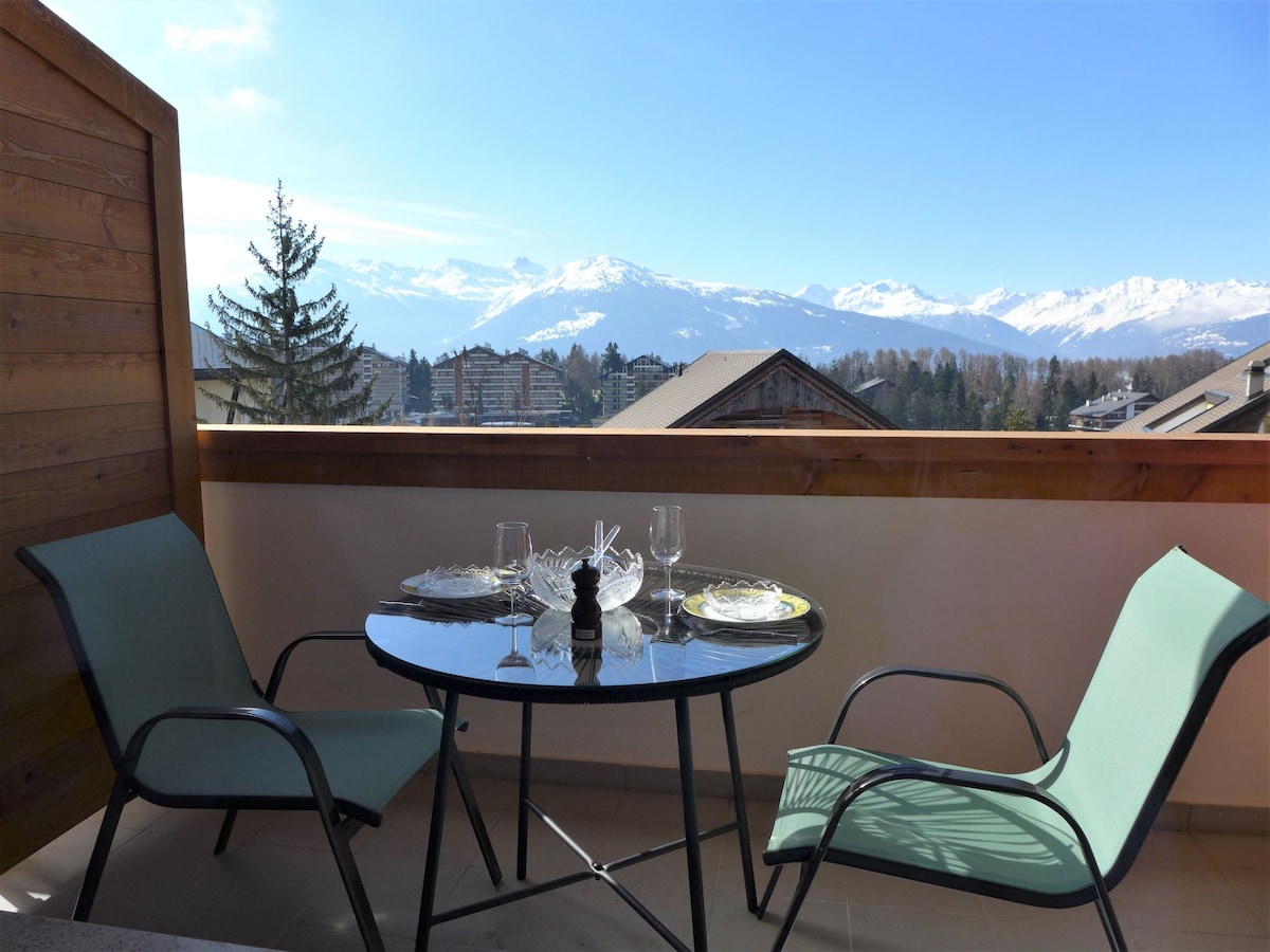 A balcony is shown with two green chairs placed around a round glass table. The table is set with dinnerware, and a scenic view of mountains stretches across the background, offering a tranquil outdoor space.