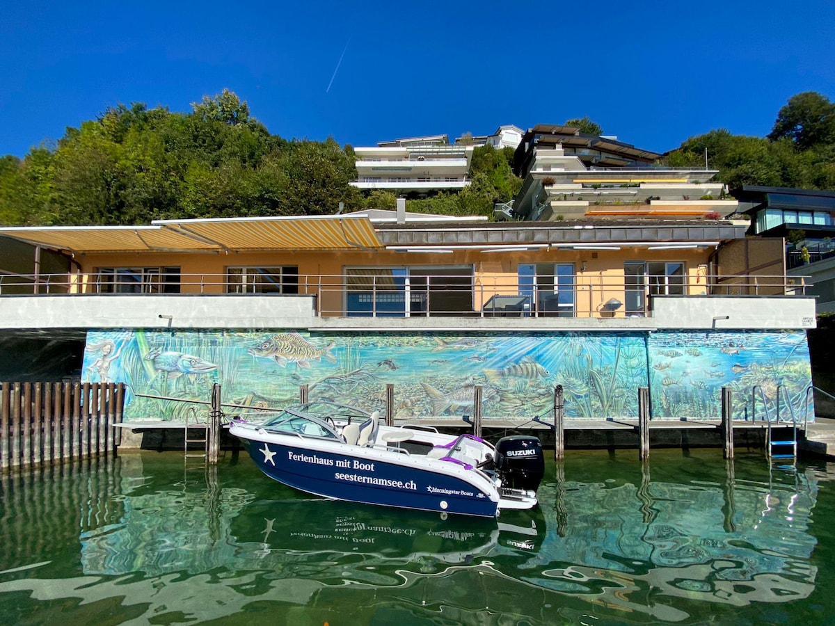 The house is positioned alongside the lake, featuring a large terrace above the water and a mural depicting aquatic life. A motorboat is docked in front, with clear water reflecting the warm sunlight. The surrounding greenery complements the modern architecture of the building.