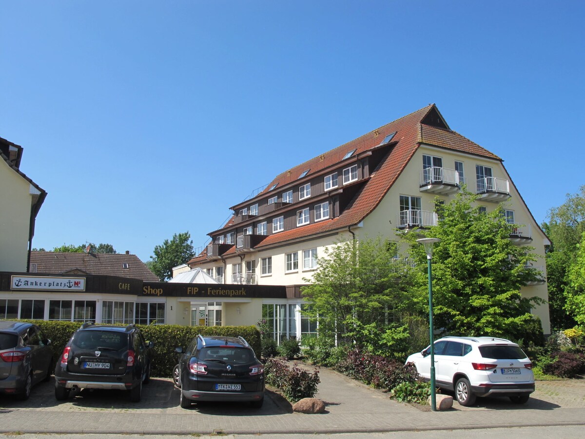 The exterior view of the FIP-Ferienpark features a three-story building with a sloped roof. Surrounding greenery is visible, alongside a parking area with several cars. A shop entrance is positioned in the foreground, and clear blue skies fill the background.