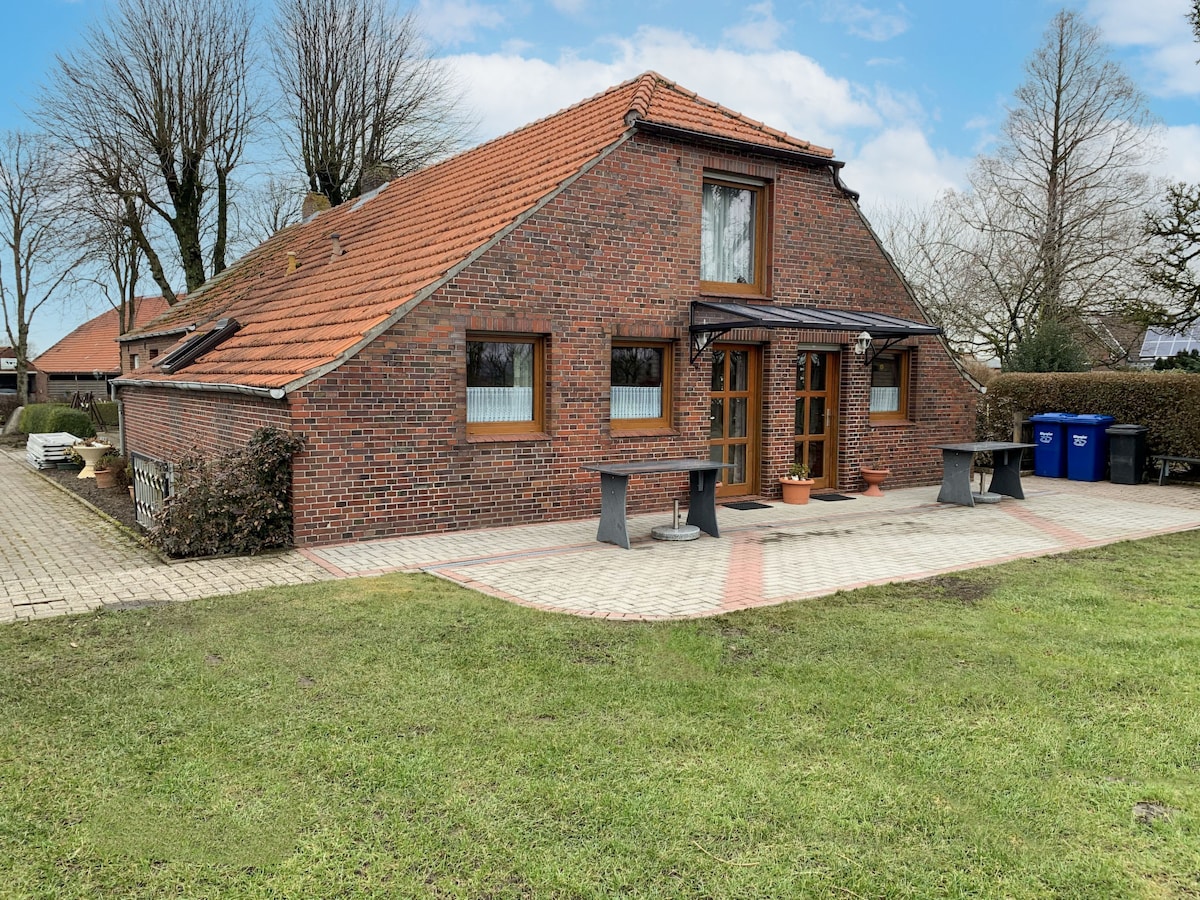 The exterior of the house features a charming brick facade with a sloped roof. Large windows allow natural light to enter, while a paved area in front provides space for seating. A well-maintained lawn surrounds the property, enhancing the tranquil setting.