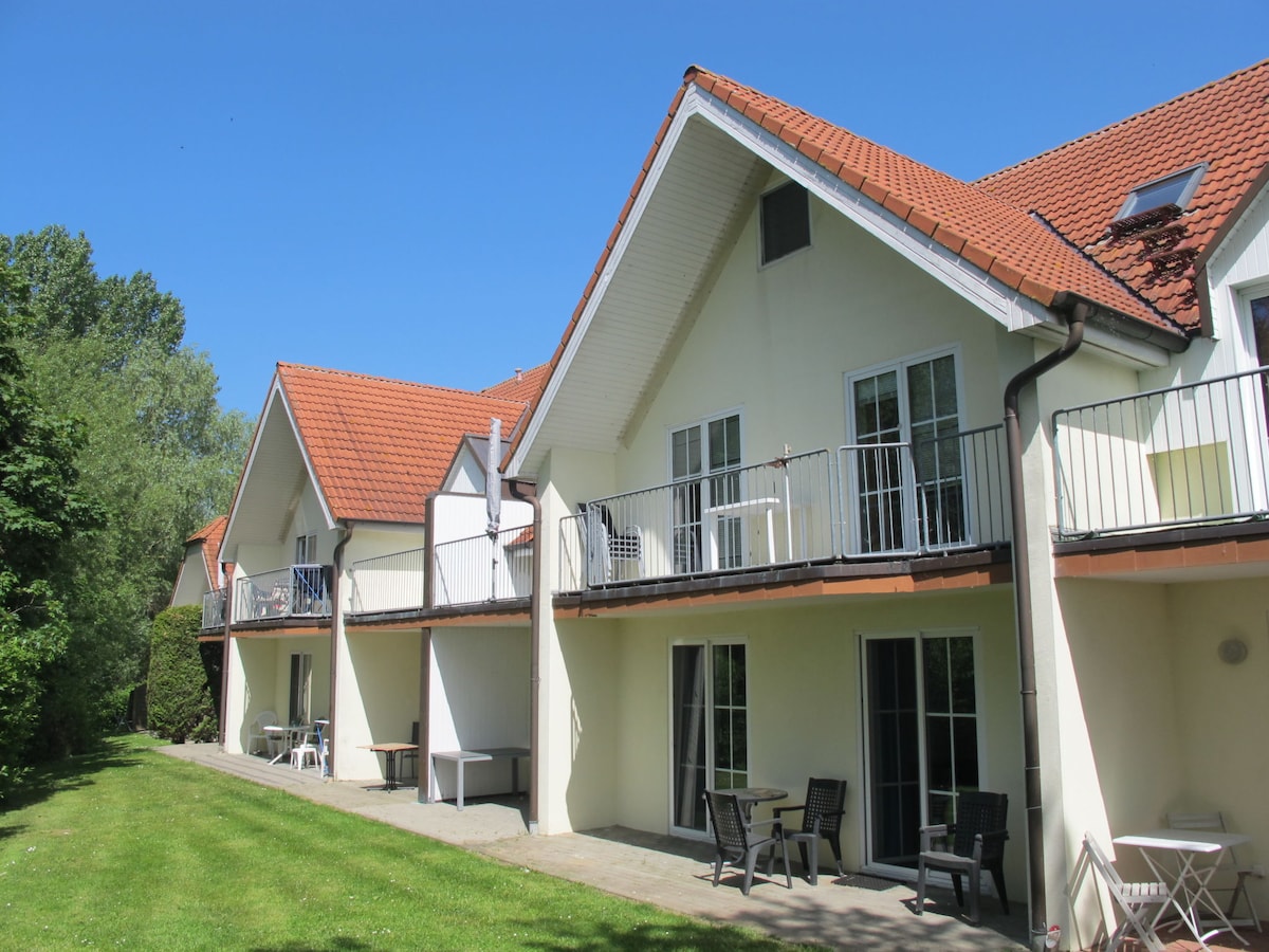 Two adjoining two-storey buildings are shown, with balconies accessible from the upper floors. The exterior features a bright, light-colored facade and a terracotta tiled roof. A lush green lawn extends in front, contributing to the scenic, natural environment.
