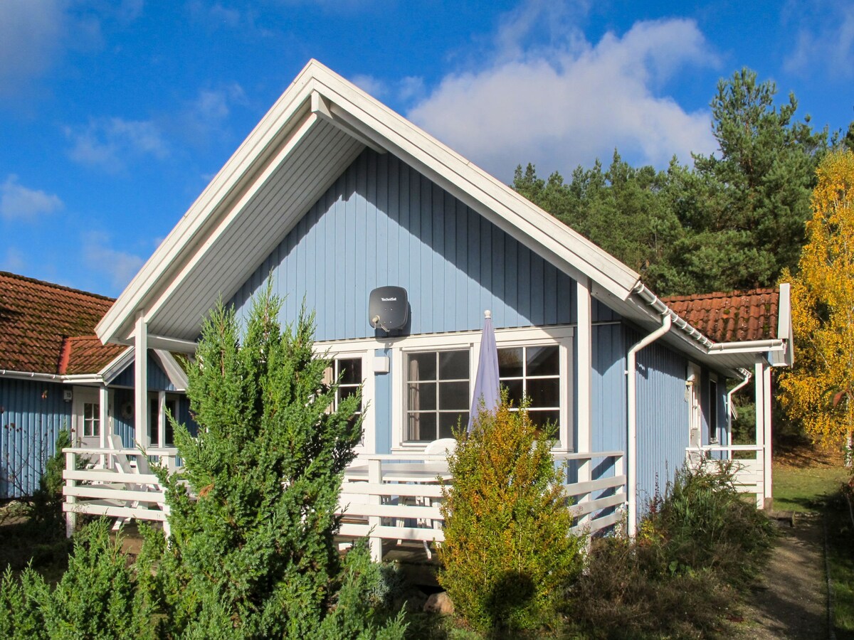 A blue house with a pitched roof is surrounded by greenery. Large windows allow natural light to fill the interior. A terrace area is visible at the front, partially shaded by a nearby tree. The exterior walls are complemented by white trim.