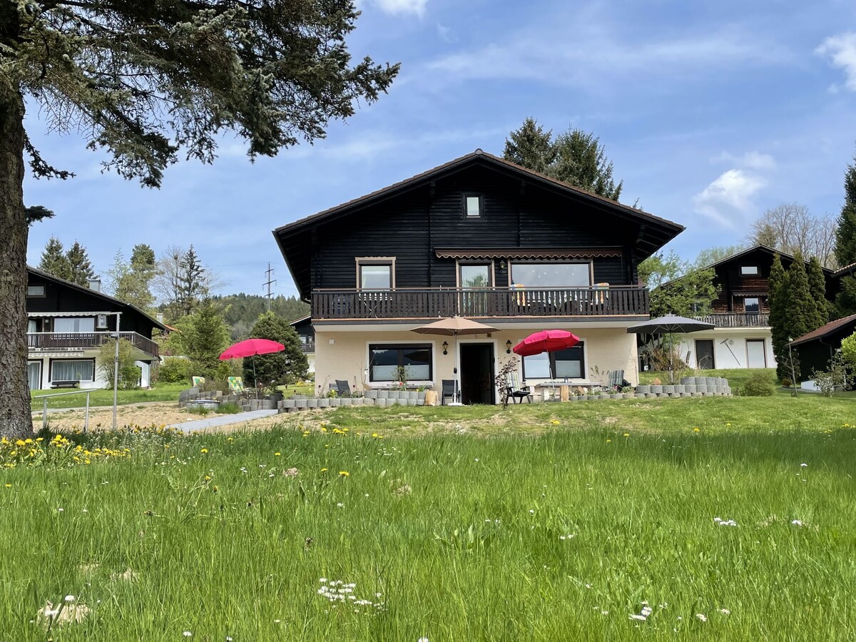 A view of the two-story building surrounded by lush green grass and colorful wildflowers. The entrance is accented by two red umbrellas, enhancing the outdoor seating area. Trees provide a natural backdrop, while nearby homes can be seen in the distance under a clear blue sky.