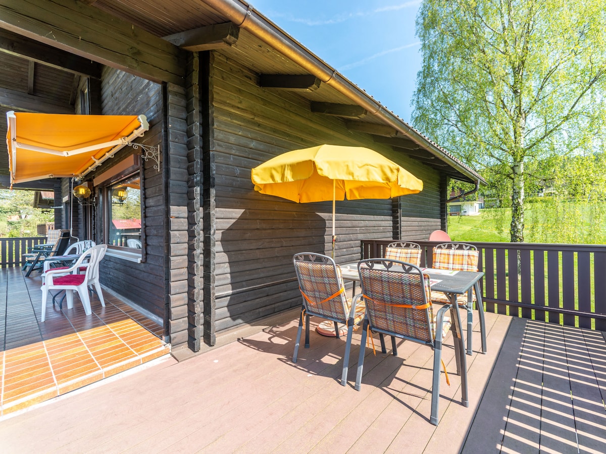 A spacious balcony features a table with chairs and a large yellow umbrella, providing shade. The balcony is surrounded by a wooden railing and offers views of a green landscape. Sunlight illuminates the area, creating a bright and inviting space.