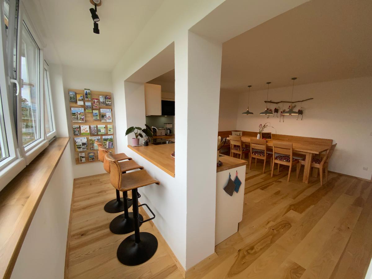 An open kitchen area features a countertop with bar stools, leading to a dining space with a large wooden table. Natural light enters through multiple windows, enhancing the bright, airy ambiance. A wall display of photographs adds a personal touch.