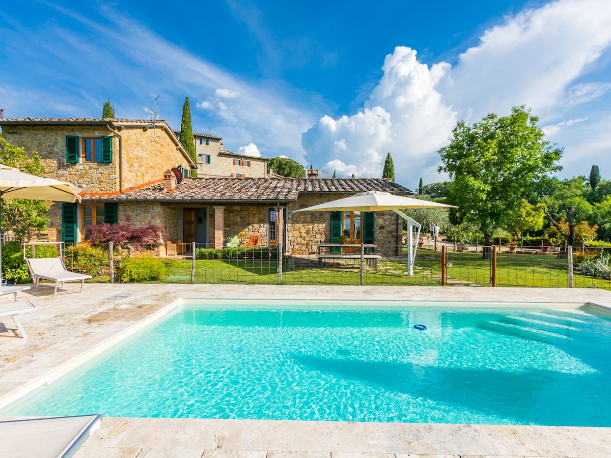 A swimming pool is set against a backdrop of a charming stone house. Lush greenery surrounds the pool area, with sun loungers positioned nearby. A shaded seating area is visible, providing a serene environment, while cumulus clouds drift across a bright blue sky.