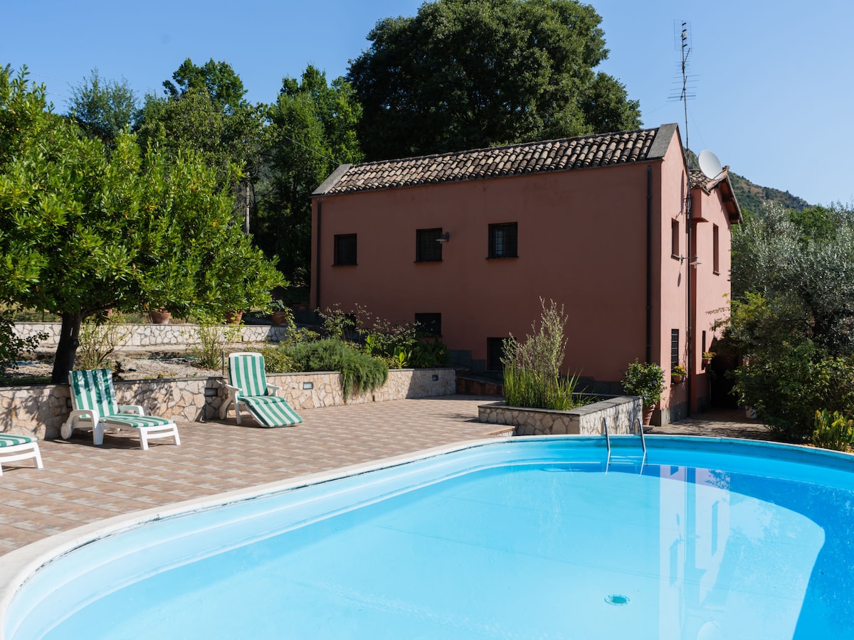 A tranquil outdoor setting features a swimming pool with clear blue water, surrounded by tiled flooring and lounge chairs. The pink villa, complemented by lush greenery, is positioned in the background, enhancing the peaceful ambiance. Trees provide shade and natural elements to the scene.
