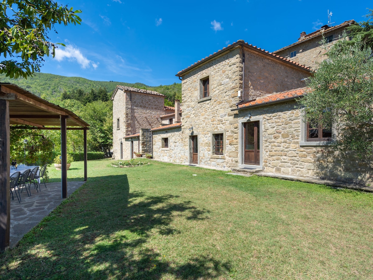 The exterior of the rustic stone house is set against a clear blue sky, with a well-kept lawn in the foreground. A covered terrace with seating is present, surrounded by greenery. The yard opens up to a scenic view of the surrounding hills.