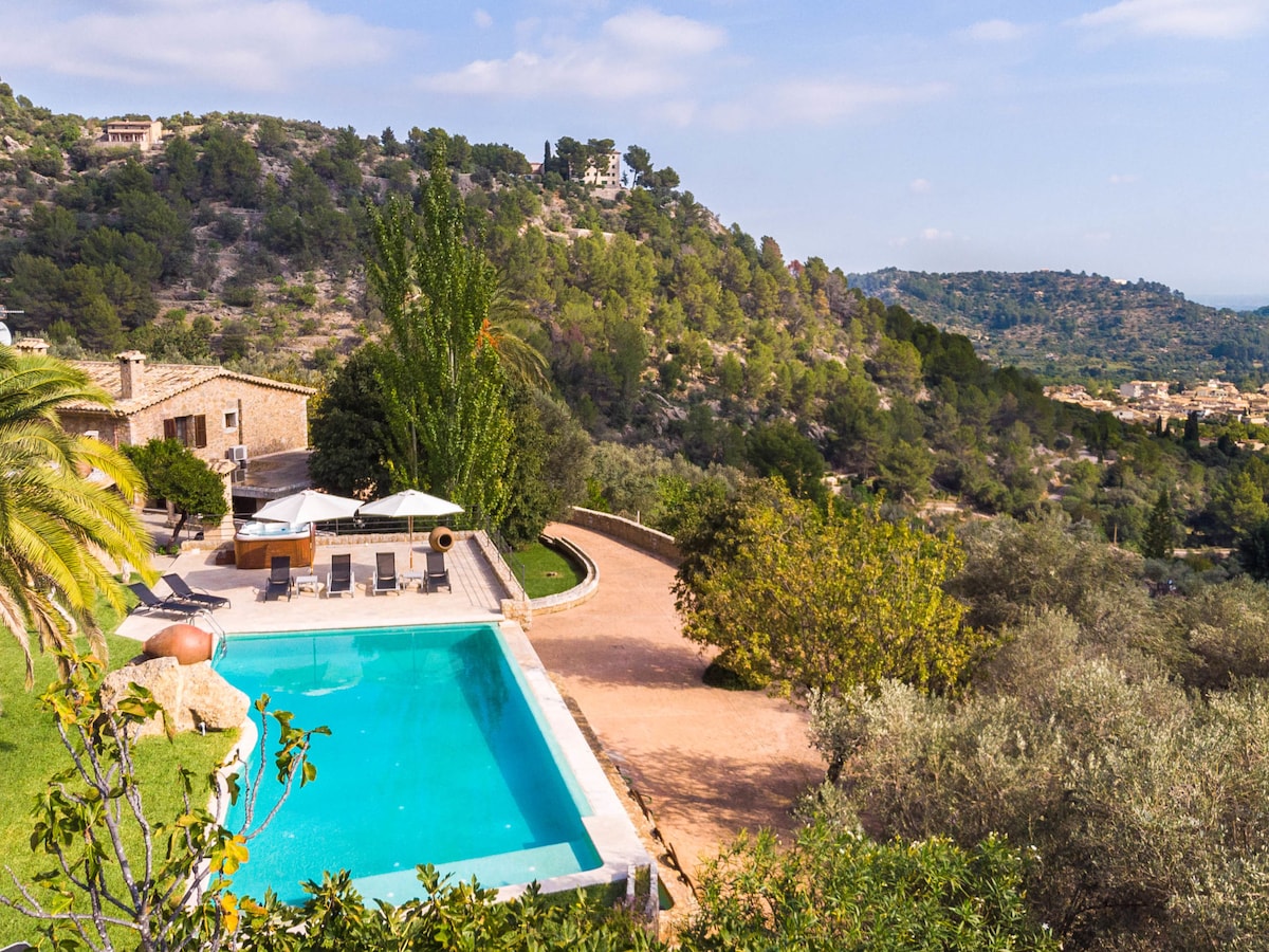 A spacious outdoor area features a rectangular swimming pool surrounded by landscaped gardens. Sun loungers are positioned nearby while the verdant hills create a scenic backdrop. The patio area offers ample space for relaxation amidst a tranquil environment.