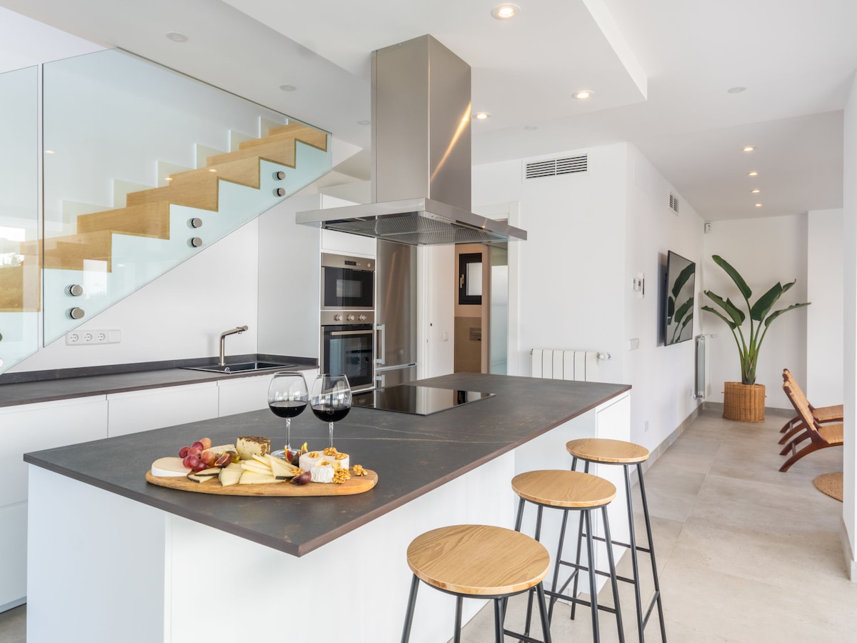 An open kitchen is highlighted by a central island with seating, showcasing a platter of cheese and fruit. Modern appliances are visible, along with a stylish staircase in the background. Natural light illuminates the space through large windows.