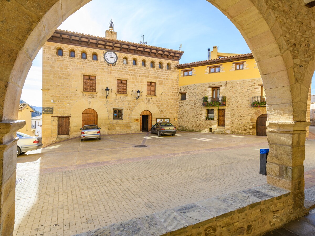 An arched stone passage frames a view of a quaint town square featuring historic buildings. A clock adorns the stone facade of one building, while parked cars line the cobblestone street. Rustic charm is emphasized by the surrounding architecture under a bright blue sky.