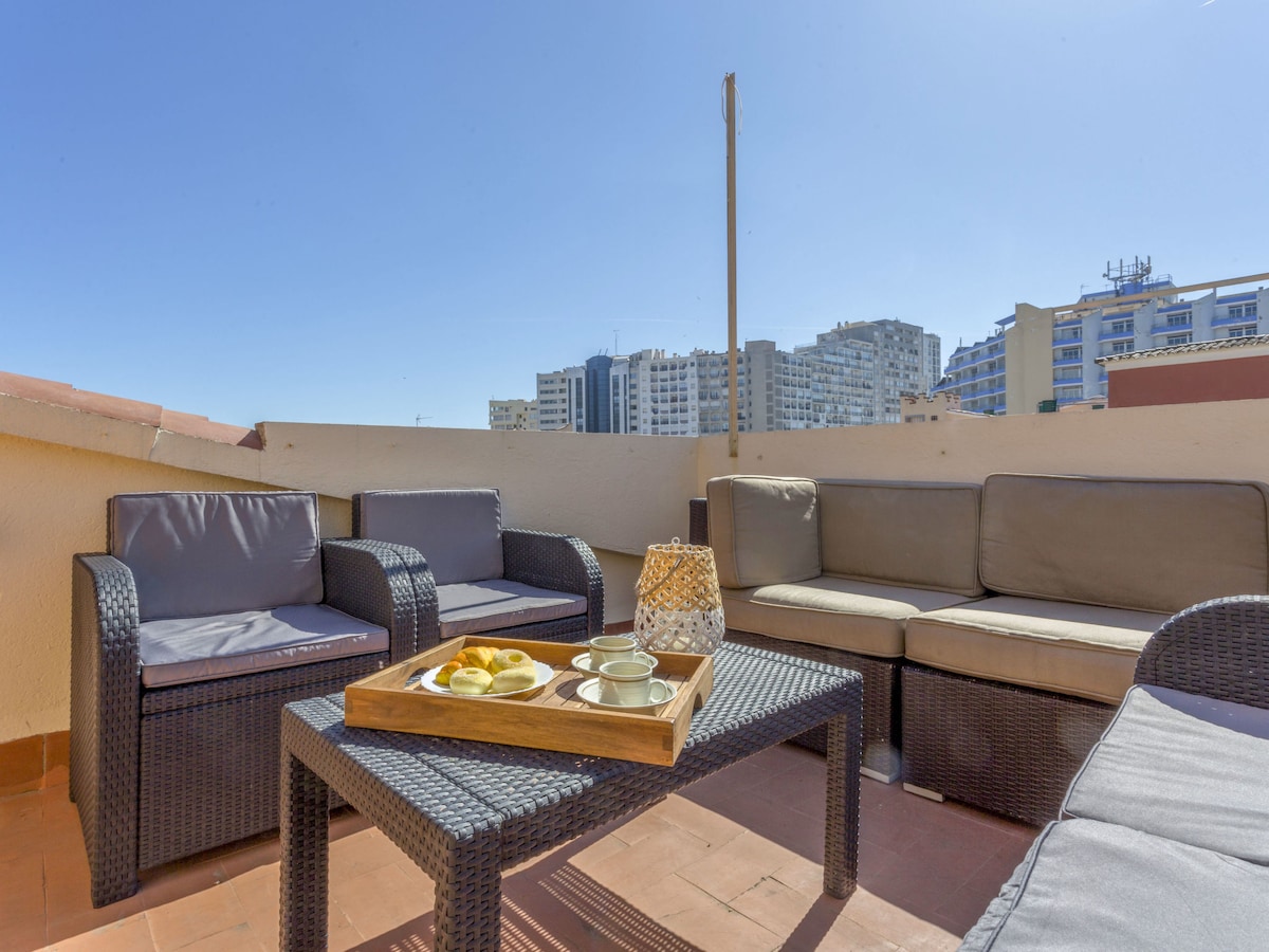 A spacious terrace area is furnished with a sectional outdoor sofa and a coffee table. A tray with snacks is placed on the table, inviting relaxation. The backdrop features buildings under clear blue skies, creating a sunny and pleasant environment.