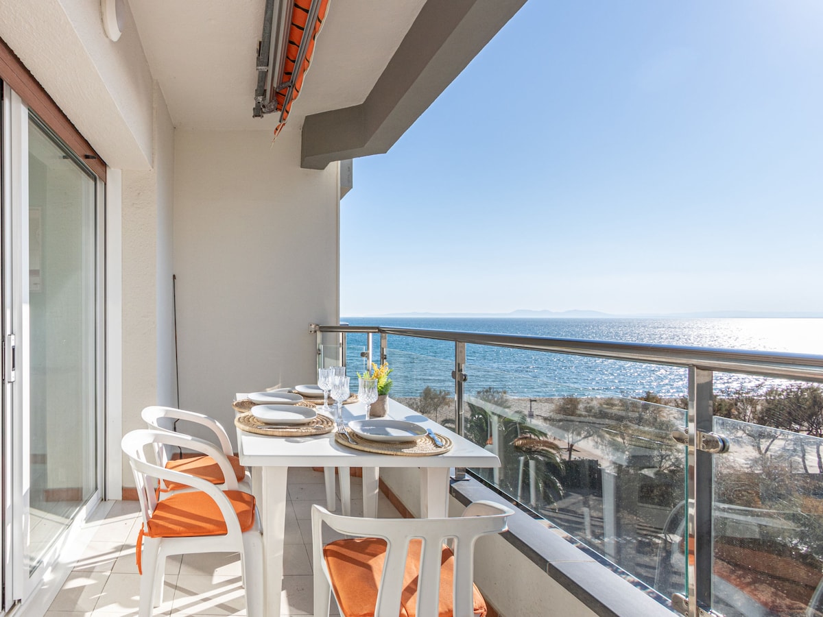 A spacious terrace is depicted, featuring a dining table set with tableware and surrounded by four white chairs with orange cushions. The railing offers a clear view of the sea, with sunlight reflecting on the water's surface.