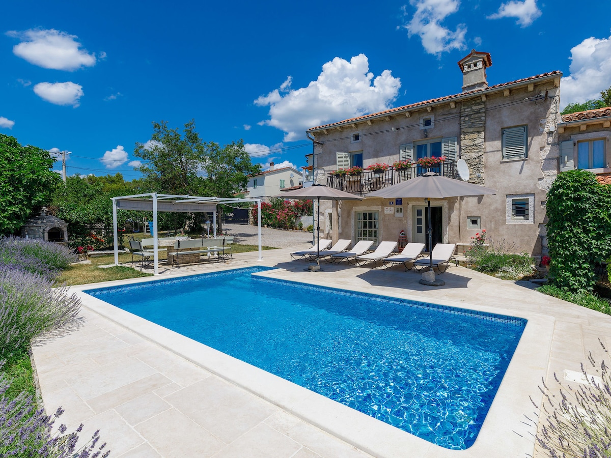 A serene outdoor space features a swimming pool surrounded by a spacious patio. Sun loungers are arranged under shaded areas with umbrellas. The house, showcasing rustic architecture, stands in the background amidst greenery and blooming flowers, enhancing the tranquil environment.