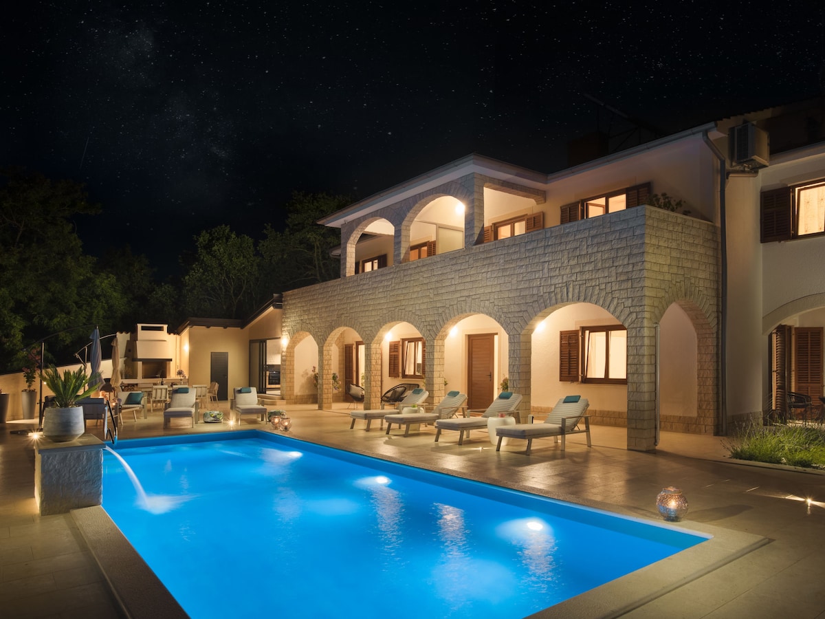 The two-story villa is illuminated under a starry sky, featuring an inviting swimming pool surrounded by lounge chairs. Arched doorways open to the terrace, showcasing warm light from the windows, while lush greenery provides a serene backdrop.