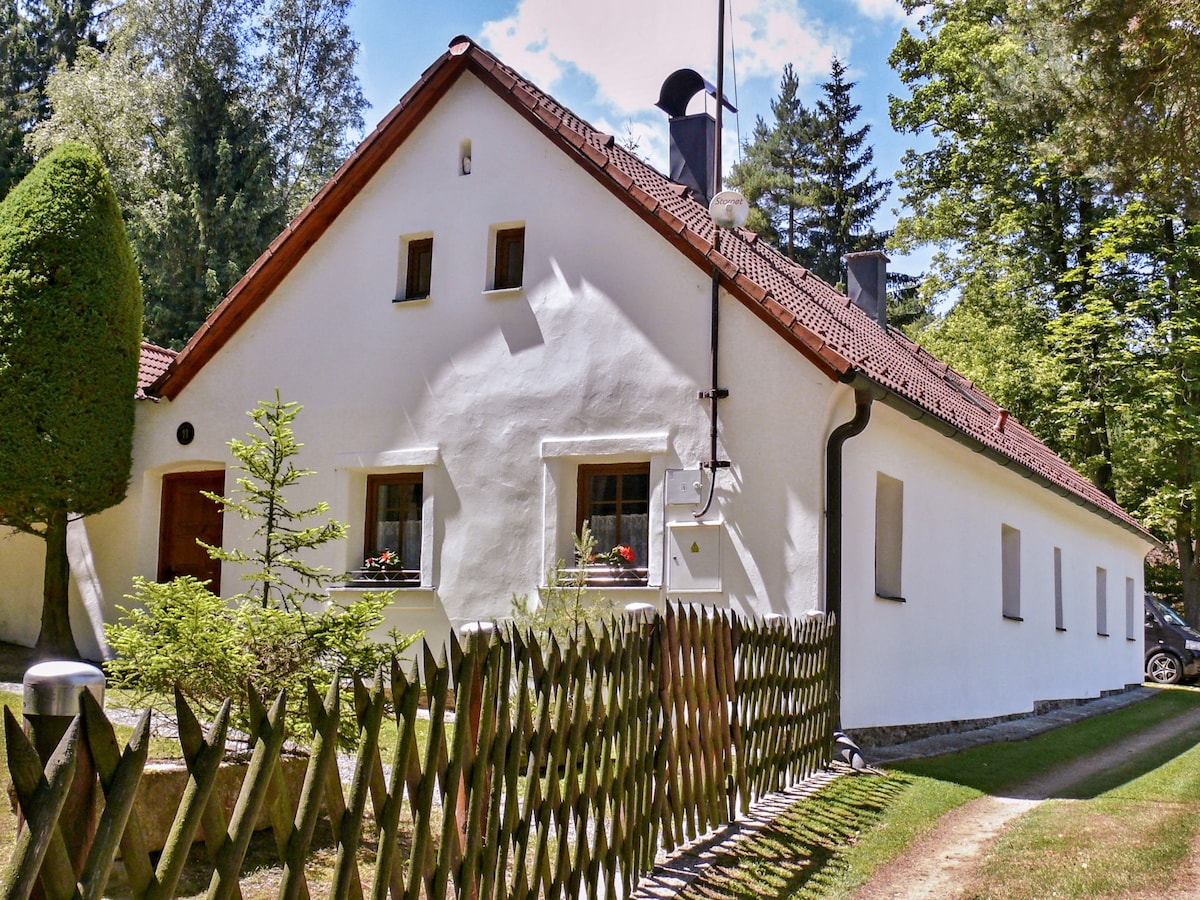 A charming country house is seen featuring a smooth white exterior and a sloped red-tiled roof. Windows adorned with planters can be spotted. A wooden fence frames the property, while lush greenery and trees provide a natural backdrop.