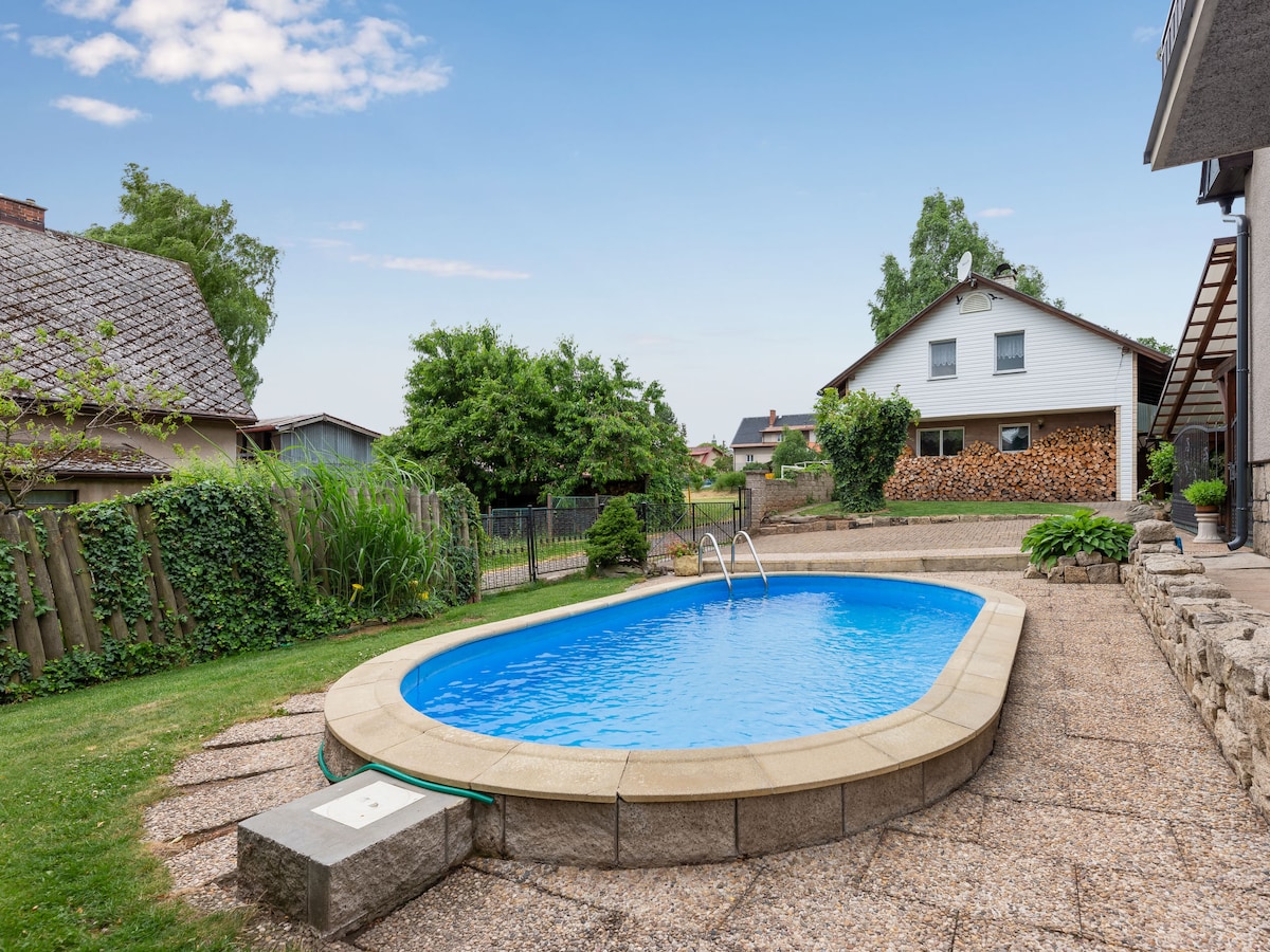 A sunken swimming pool is featured in a private outdoor space, surrounded by well-maintained grass and stone pathways. Lush greenery and wooden fencing provide a natural backdrop, while a cozy house structure can be seen in the distance.