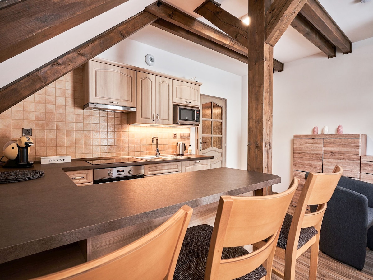The kitchenette features wooden cabinetry and modern appliances, including a microwave and kettle. A bar area with four wooden stools invites casual dining. Exposed wooden beams add architectural interest, and soft lighting highlights the functional workspace.