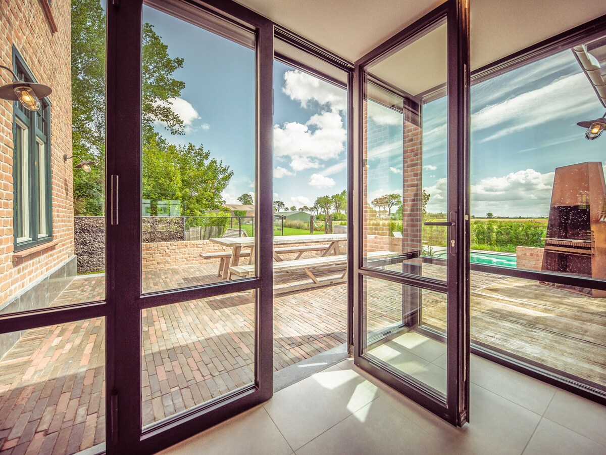 Large glass doors frame a view of the outdoor patio and swimming pool, inviting natural light into the interior. A wooden dining table is positioned outside, complemented by a serene landscape of greenery and blue skies, creating a connection between indoor and outdoor spaces.
