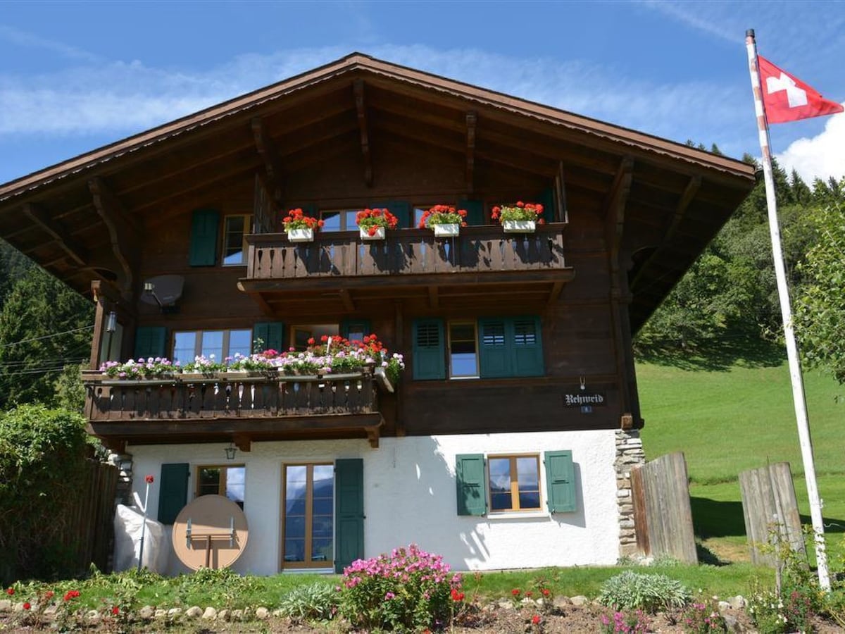 A traditional Swiss chalet is showcased, featuring a sloped wooden roof and a bright Swiss flag. Flower-filled balconies add color, while the lower level presents a white facade with green shutters. A well-maintained garden with flowers enhances the property's appeal.