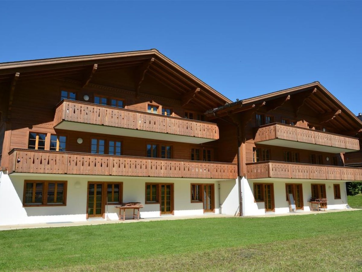 A spacious, wooden building with multiple balconies overlooks a green lawn. The design features large windows and a charming balcony railing. The clear blue sky above enhances the welcoming atmosphere of the property.