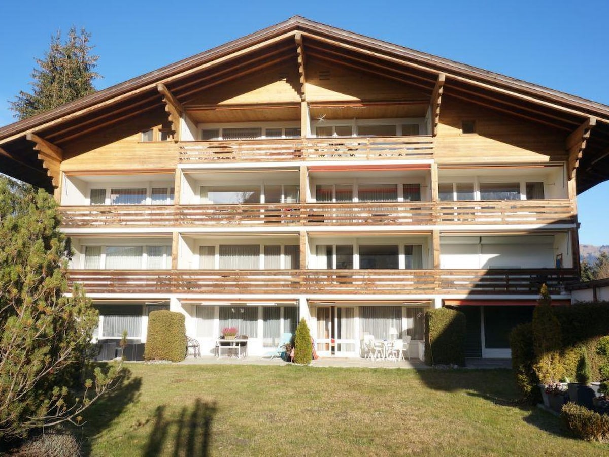 The apartment building features a rustic wooden facade with multiple balconies adorned with wooden railings. A well-manicured lawn in front is complemented by shrubs, and the structure is set against a clear blue sky, indicating a bright and sunny location.