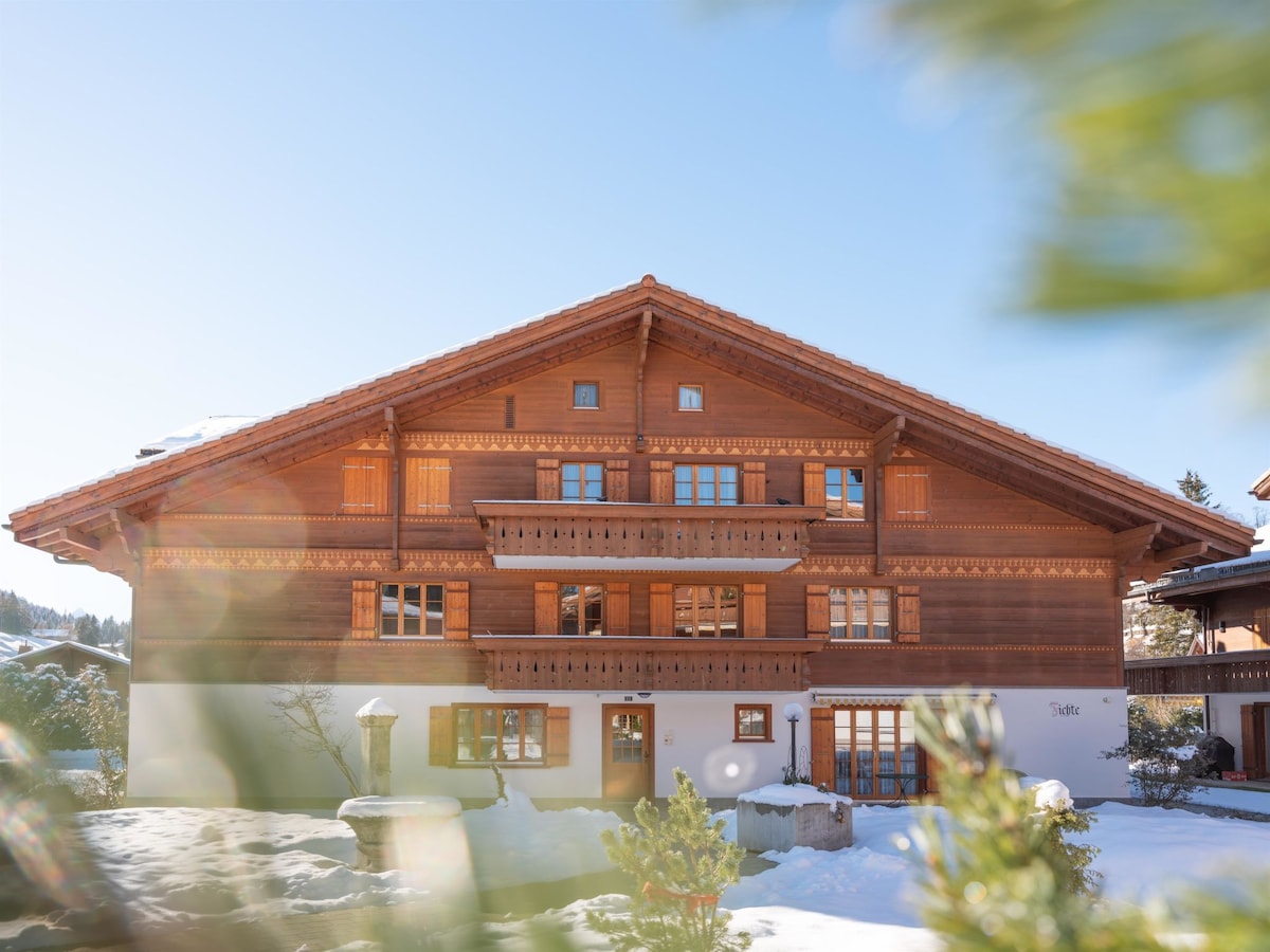 A multi-family chalet is depicted, showcasing traditional wooden architecture with decorative carvings. Multiple windows and balconies are featured on the facade, surrounded by a snow-covered landscape. The clear blue sky creates a bright backdrop for this well-maintained property.