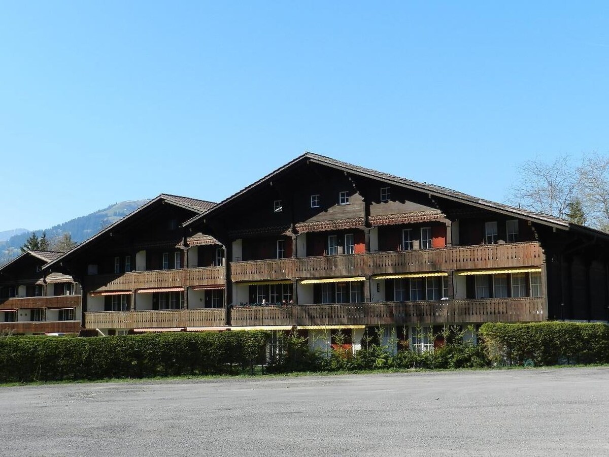 A three-story wooden building features numerous balconies adorned with flower boxes. The structure is positioned against a backdrop of hills, contributing to the serene environment. Ample outdoor space is visible in the foreground, with a clear blue sky overhead.