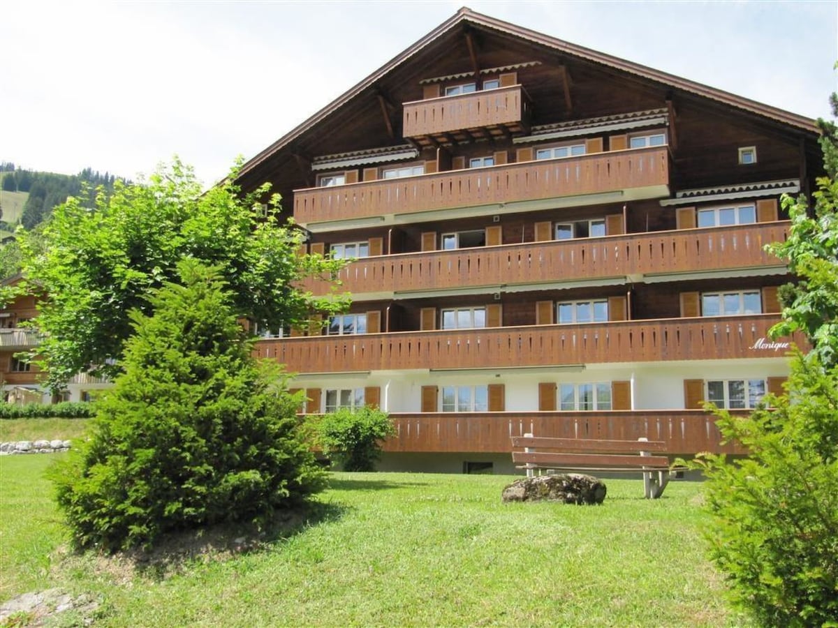 The exterior of a three-story wooden apartment building is surrounded by well-maintained greenery, featuring multiple balconies with wooden railings. A grassy area with seating is visible in front, offering a peaceful setting with trees enhancing the natural ambiance.