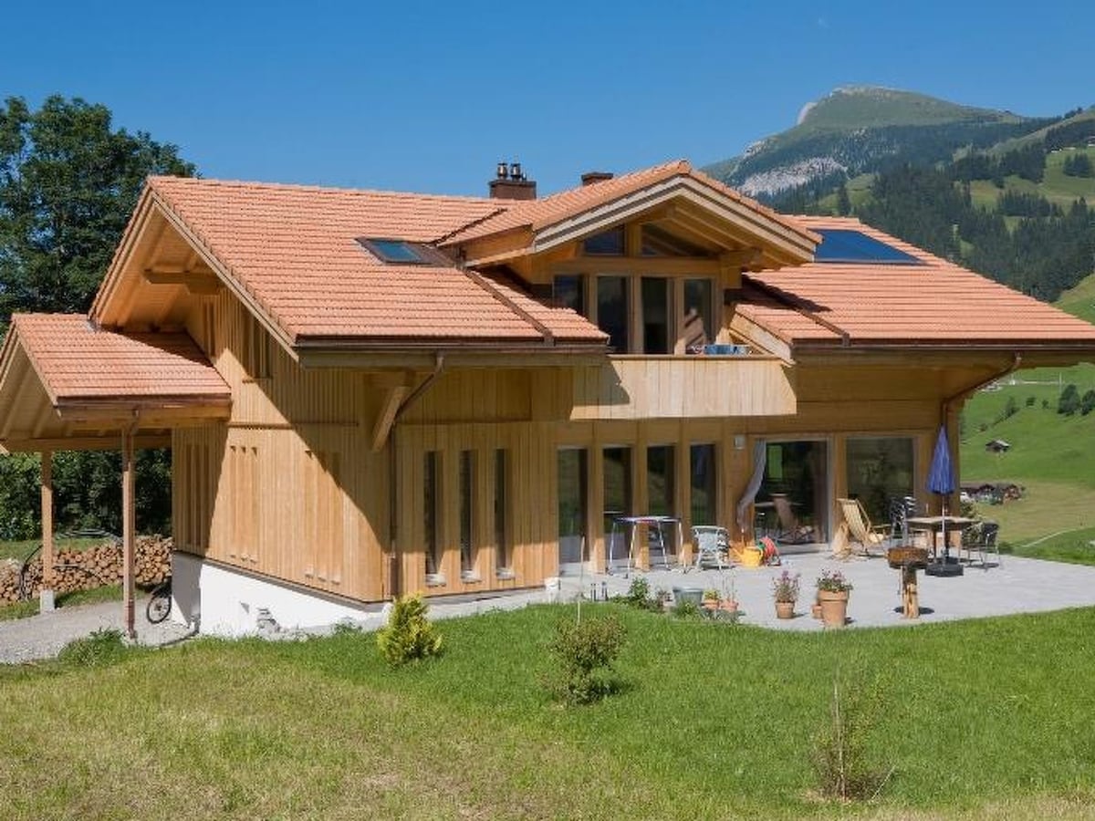 A modern two-story wooden house is set against a backdrop of rolling hills and clear blue skies. Large windows and a covered patio are visible, along with potted plants and outdoor seating. The sloped roof features skylights, enhancing natural light throughout the space.