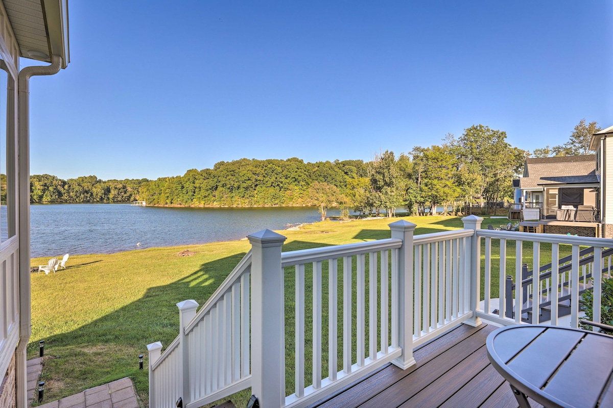 A scenic view of Tims Ford Lake is presented from a furnished patio. The expansive lawn leads directly to the water's edge, framed by greenery and wooded areas. White railings provide a boundary, enhancing the outdoor living space with a peaceful lakeside atmosphere.
