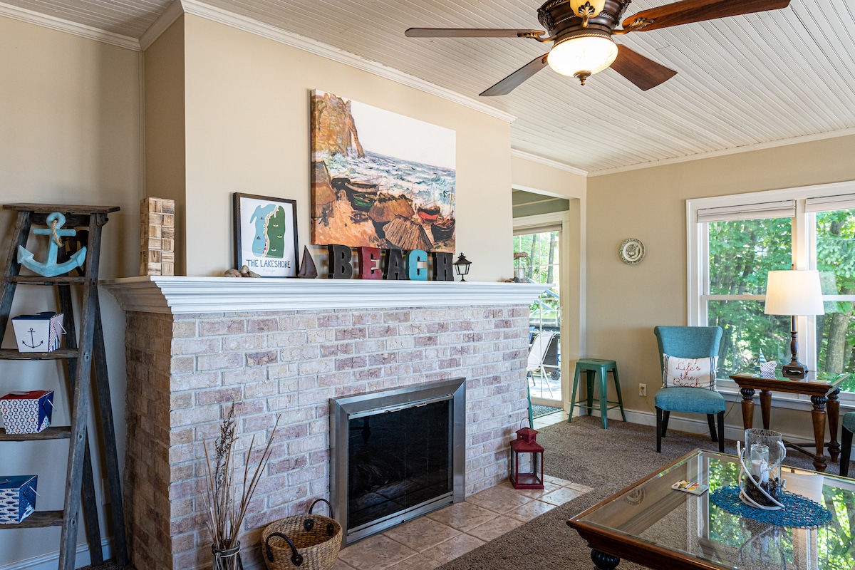 A spacious living area features a neutral color palette and a cozy fireplace with a stone facade. Decorative beach-themed art and accents are displayed on the mantel. Natural light fills the room through large windows, complemented by an inviting blue chair and a small table with a lamp.